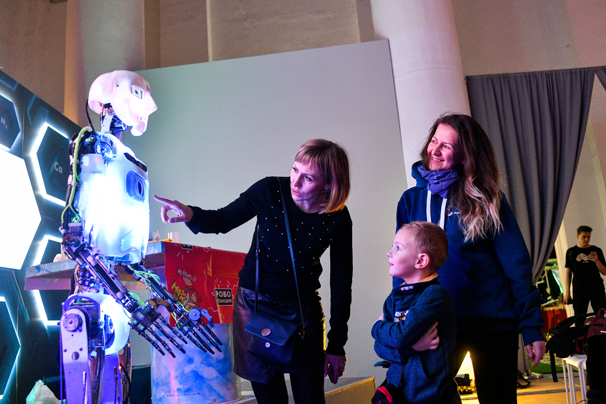 Москвичка Ядвига Елисеева (слева) показывает сыну Мише камеру, расположенную у робота Теспиана в области груди. Мальчик впервые видит такую машину вживую и внимательно слушает маму. Подруга семьи Марина Могилевцева (справа) тоже с большим интересом изучает киборга 