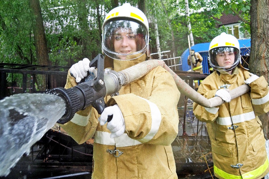 Добровольцы Дарья Львова (на переднем плане) и Елизавета Малюк в защитных костюмах показывают, как с помощью пожарного рукава тушить возгорание. Девушки прошли двухнедельное обучение во Всероссийском студенческом корпусе спасателей