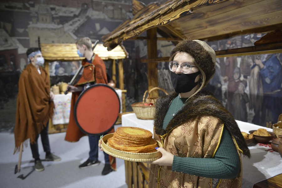 Волонтер Ирина Виноградова в старинном наряде показывает угощения, которые подавали гостям в древнерусском Новгороде и других городах. В руках участница держит убедительную бутафорию — румяные блинчики
