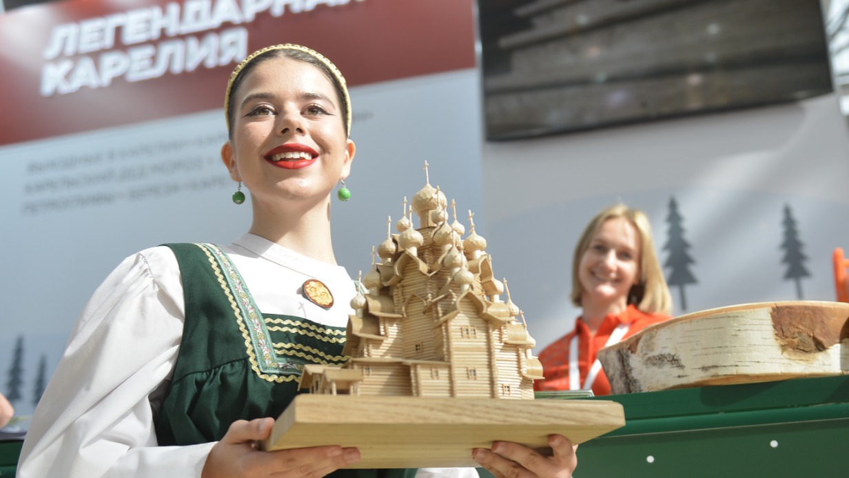 Администратор карельской базы отдыха Елена Смольская представляет сувениры от местных мастеров. На девушке надет сарафан зеленого цвета. Он символизирует природу, которой славится республика. Еще регион предлагает москвичам посетить резиденцию Деда Мороза и покататься на собачьих упряжках 