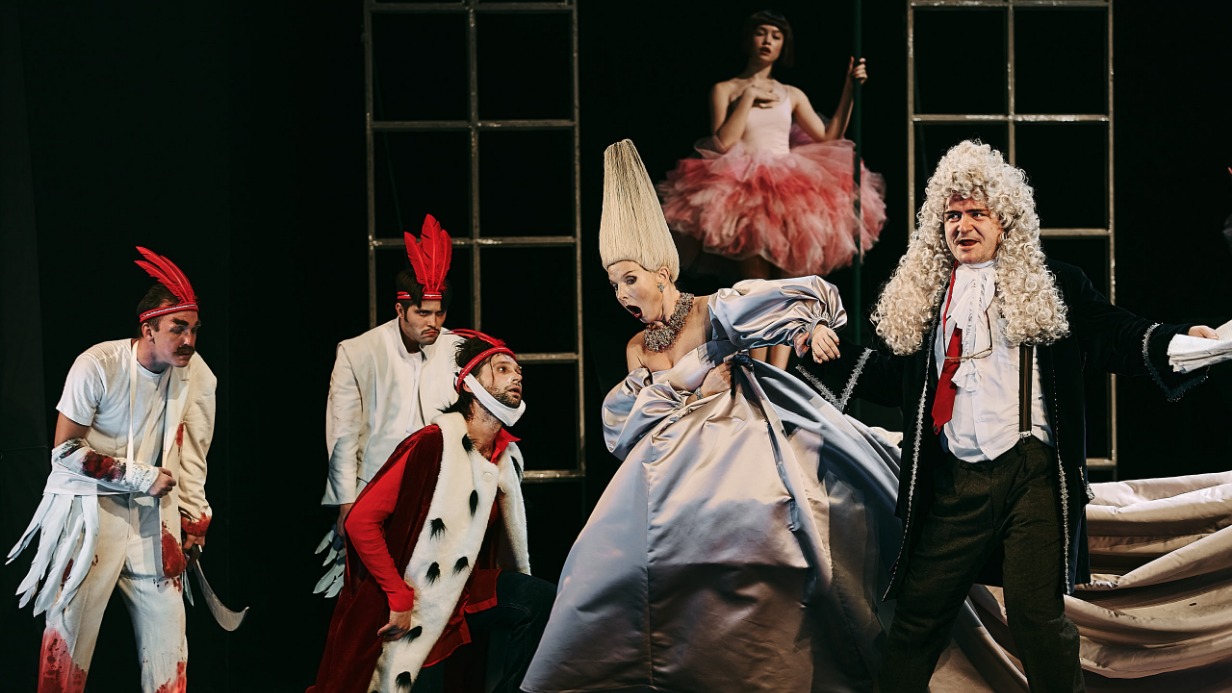 Сцена из спектакля «Багровый остров». Актер Александр Дмитриев (на переднем плане слева) в роли драматурга Дымогацкого, который согласился сыграть одного из персонажей пьесы. На сцене с ним актриса Ирина Царенко в роли Лидии Ивановны (в центре). Также в эпизоде играет актер Александр Матросов в роли Геннадия Панфиловича (справа)