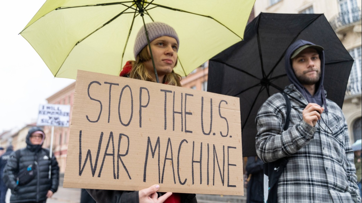 Митинг в Варшаве, на котором участник держит транспарант с надписью «Остановите США — машину войны» 